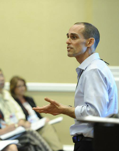 Allan Reeder speaking in front of a group, with people listening.