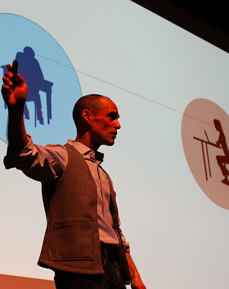 Allan Reeder holds a clicker in front of a screen displaying two people at desks.