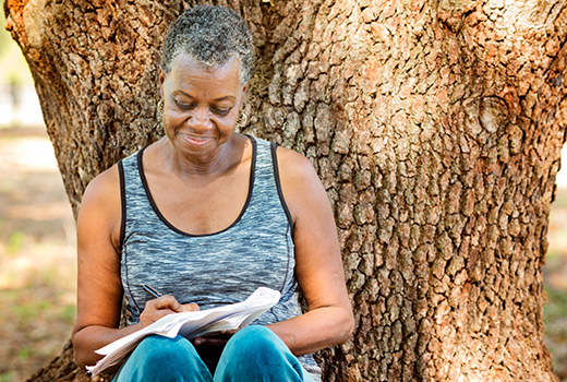 African American woman leaning against tree and writing in notebook.