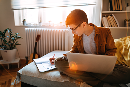 boy in room with computer and notebook, writing.