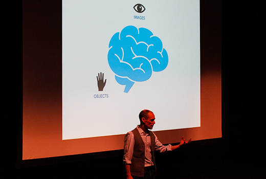 Founder Allan Reader giving a talk, standing in front of a screen with an illustration of a brain surrounded by the words: objects, images