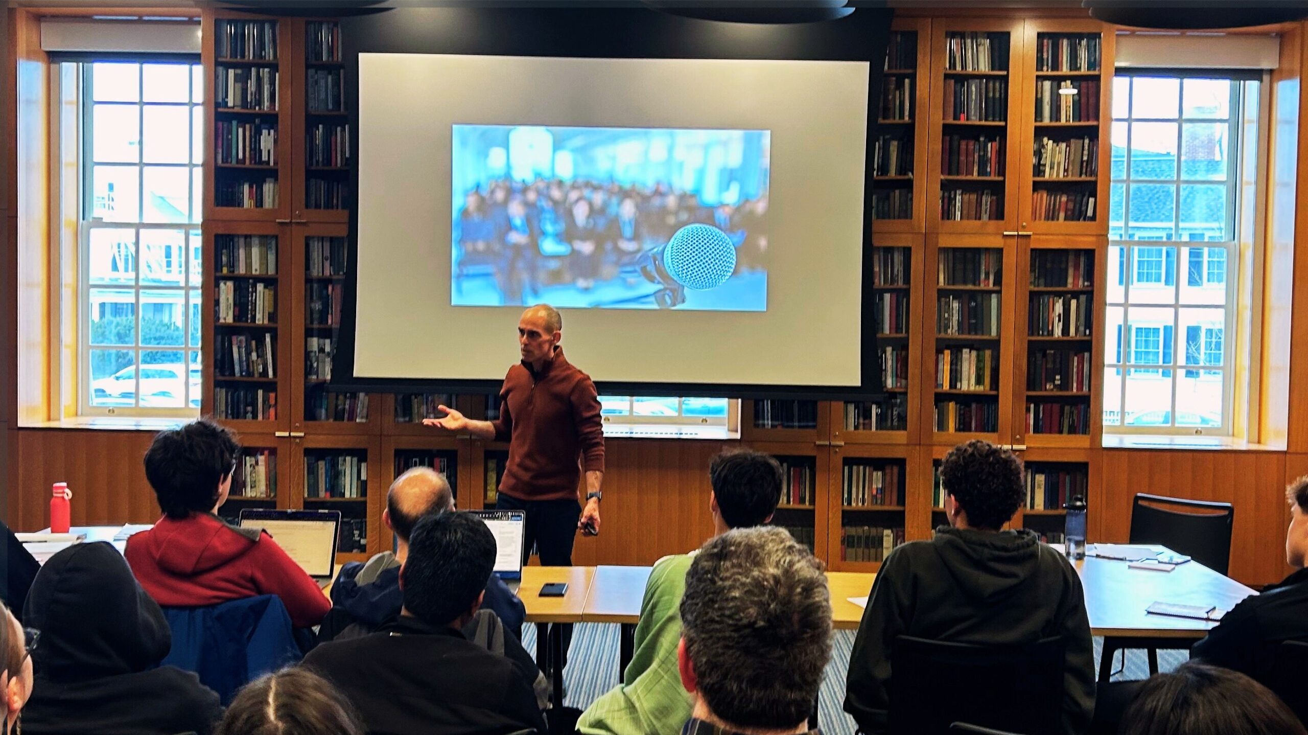 Allan Reeder teaching in a bookshelf-lined room, with an audience watching. The screen behind him shows a photo of a microphone. 